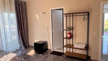 Habitación de hotel con cama, armario, un mueble negro pequeño, estantería y puerta que da a un balcón.