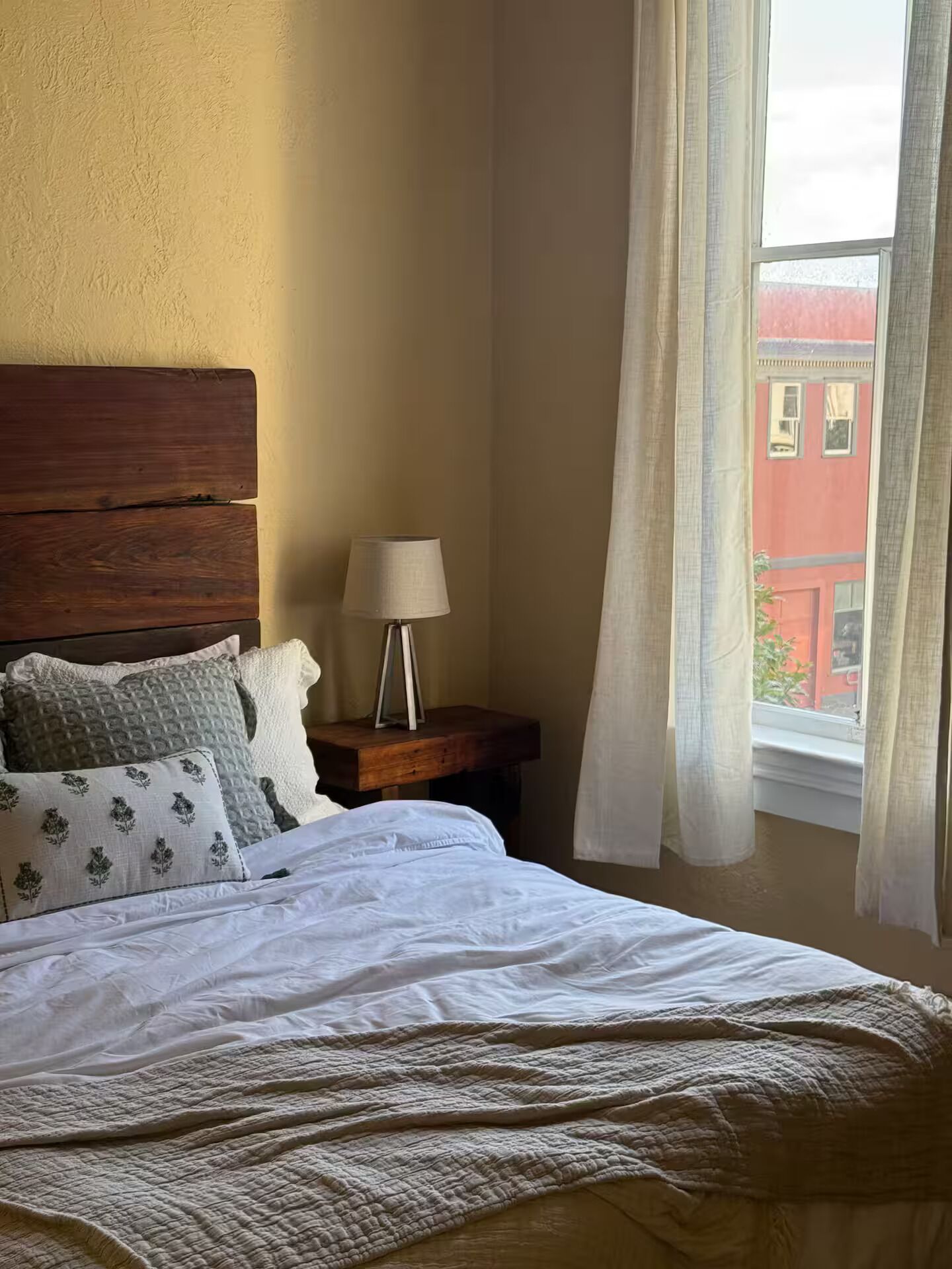A bedroom with a wooden headboard, a bedside table with a lamp, a bed with white bedding, and a window with curtains.