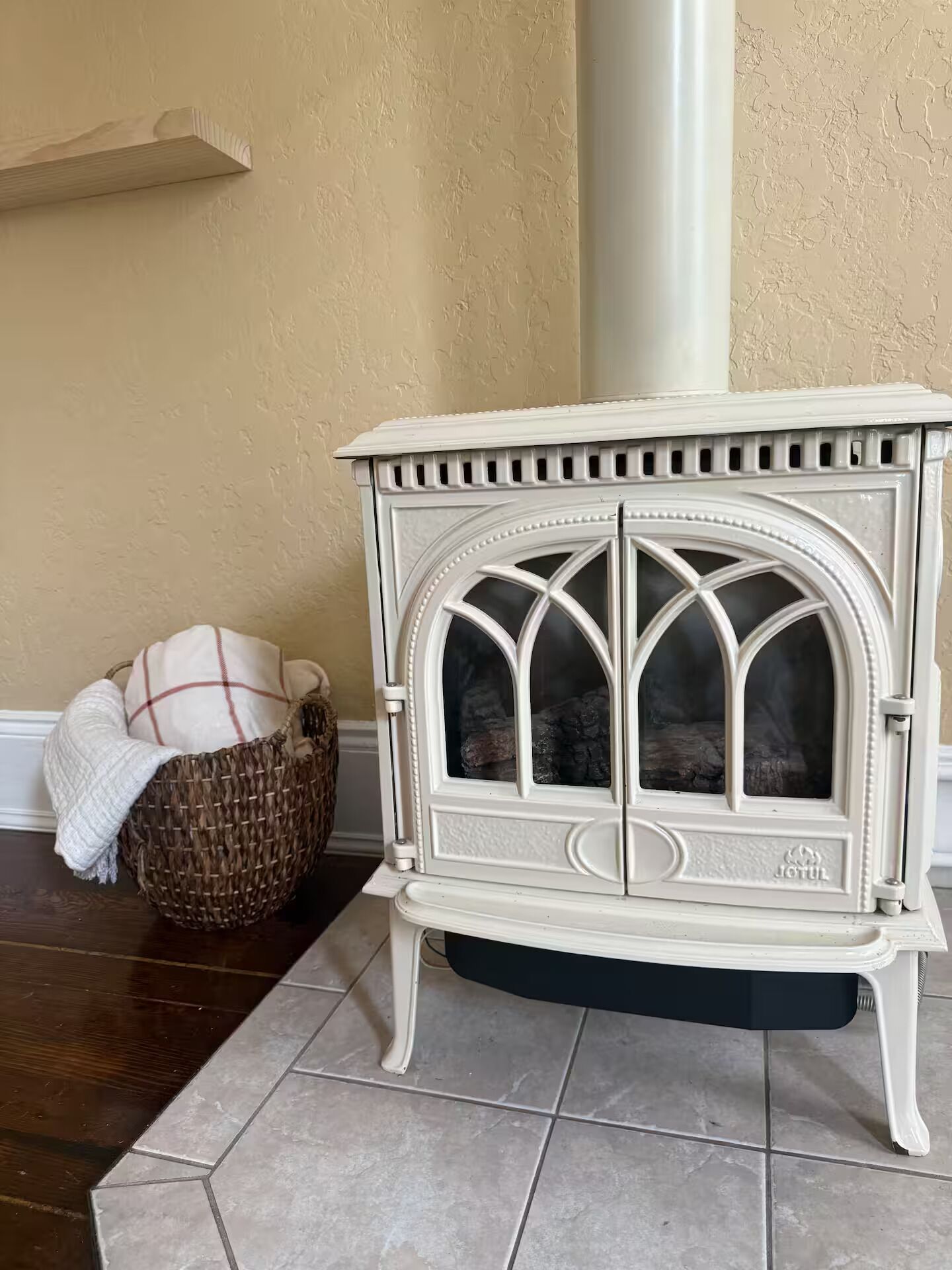 A white wood-burning stove with a glass door, positioned on a tiled floor next to a wicker basket and a white shelf.