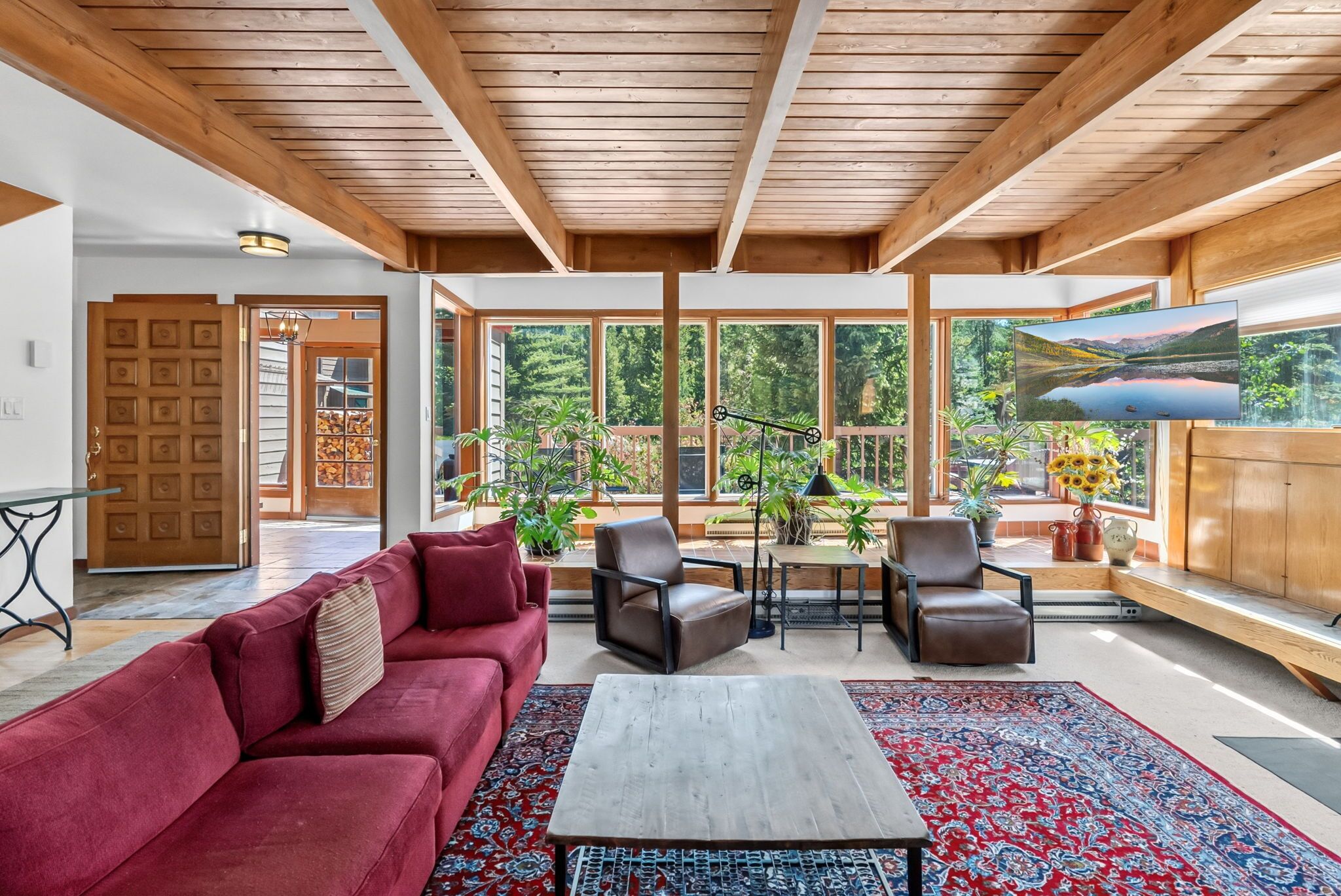 A spacious living area with a red sofa, two leather chairs, a coffee table, and a large window offering a view of greenery.