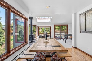 A spacious room with a large wooden table, a fireplace, and large windows offering a view of the outdoors.