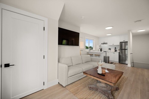 Eine moderne Wohnung mit Wohnbereich, einer Couch, einem Couchtisch, einer Küche mit weißen Schränken und einem Kühlschrank.