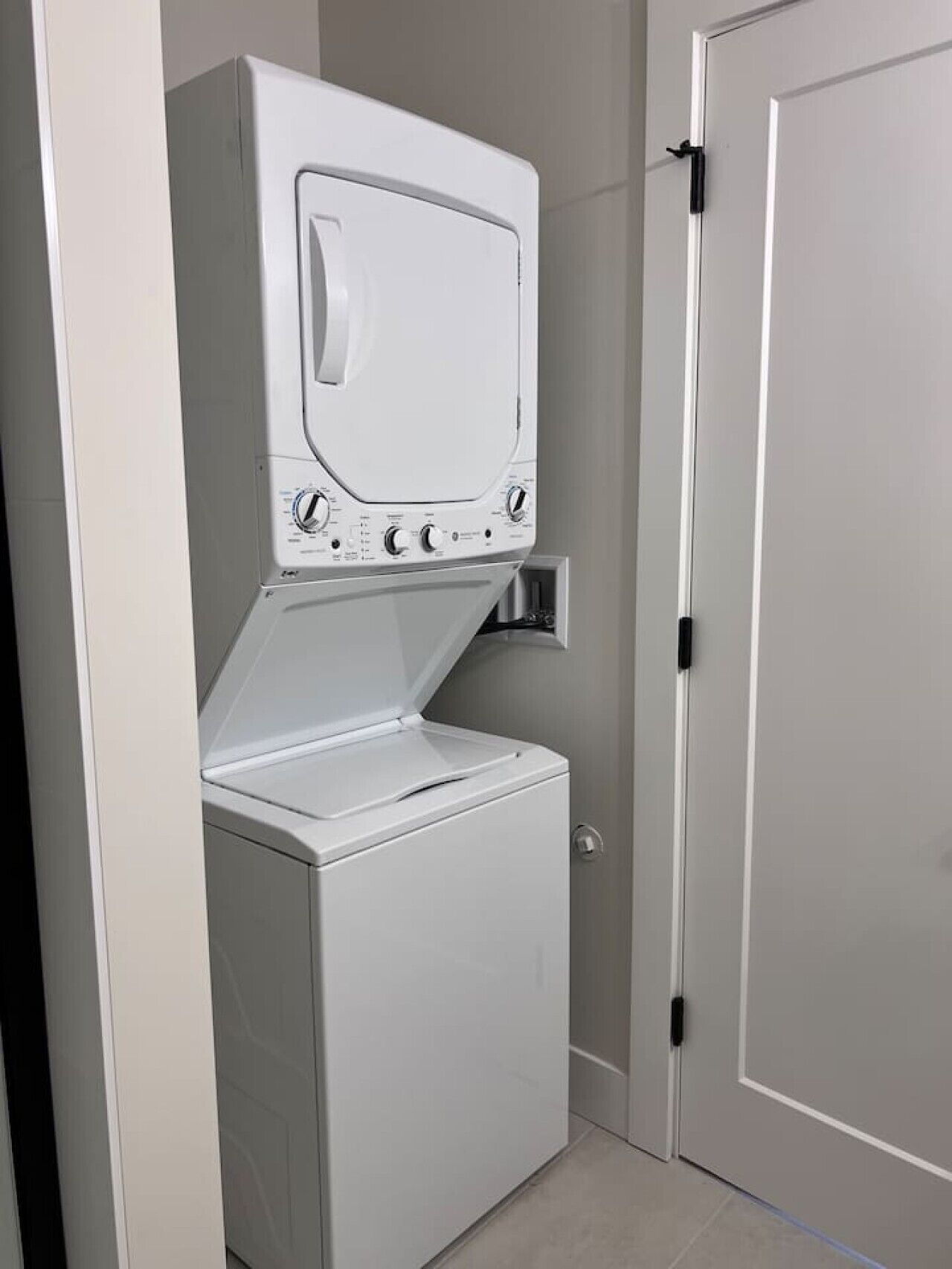A compact, white laundry room with a built-in washer and dryer, a door with a black handle, and a tiled floor.