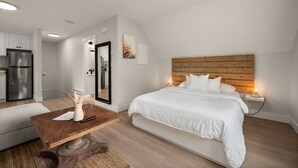 A bedroom with a wooden headboard, a bed with white bedding, a bedside table with a lamp, and a view of a kitchen with a refrigerator.