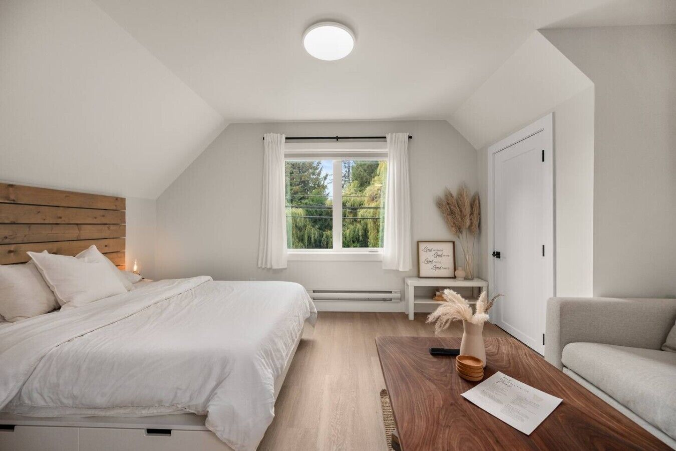A bedroom with a bed, a bedside table with a lamp, a window with curtains, and a view of greenery outside.