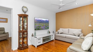 Sala de estar con piso de madera, sofá blanco, televisor montado en la pared y aparador de madera con puertas de vidrio.