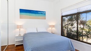 Un dormitorio con una cama, dos mesitas de noche, un ventanal con vistas a árboles y un cuadro de una playa.
