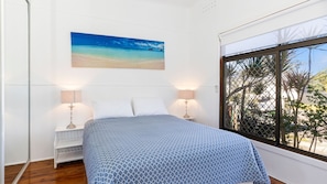 A bedroom with a bed, two bedside tables, a window with a view of trees, and a painting of a beach. - Private Oasis with pool, BBQ & entertaining areas (The Entrance)