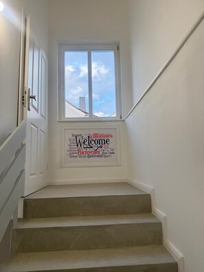 A staircase with a 'Welcome' sign in multiple languages, a window with a view of the sky and a building, and a door with a handle.