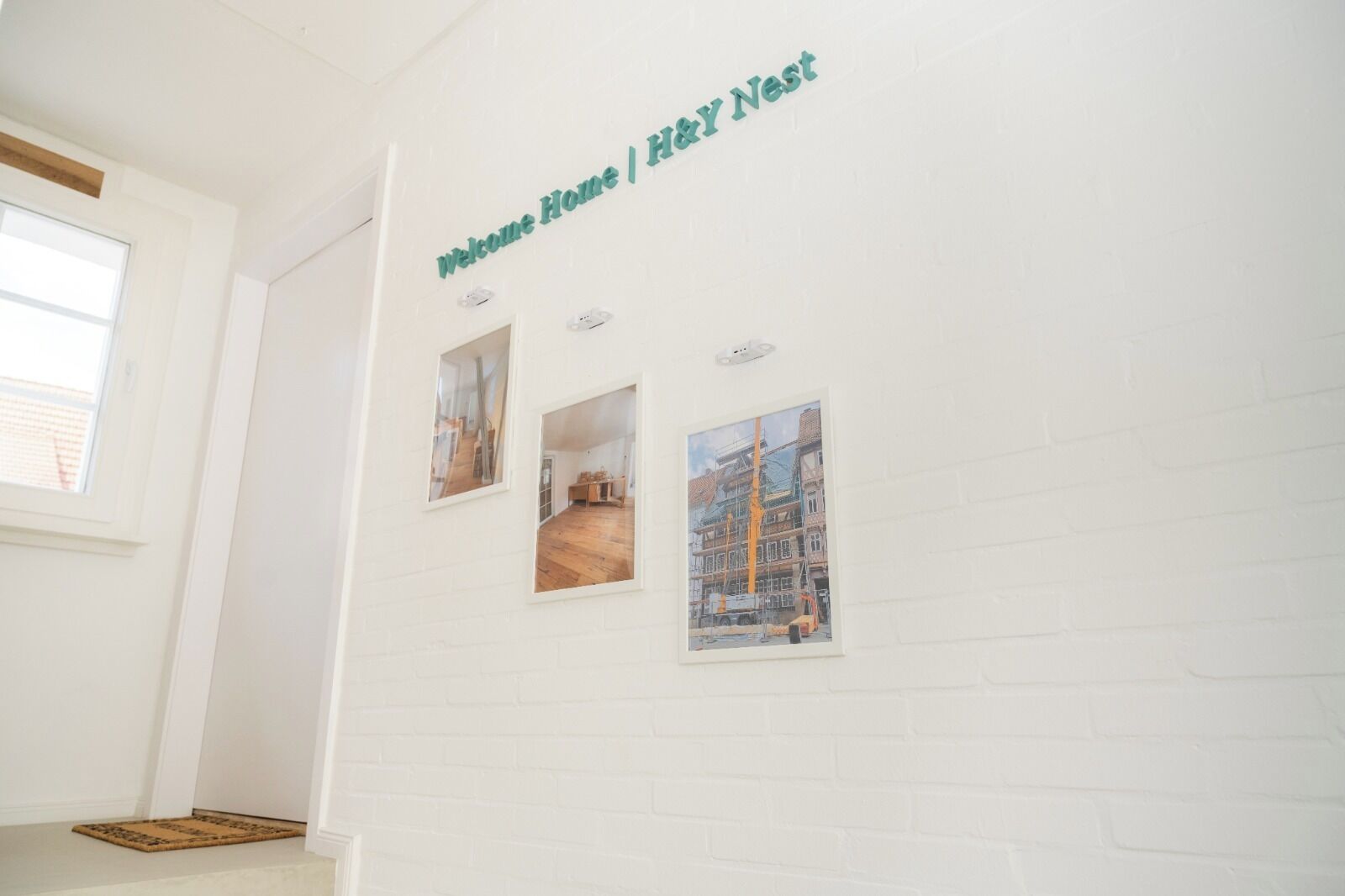 Un mur en briques blanches orné de trois tableaux encadrés et d’une pancarte indiquant « Bienvenue à la maison | H&Y Nest ».