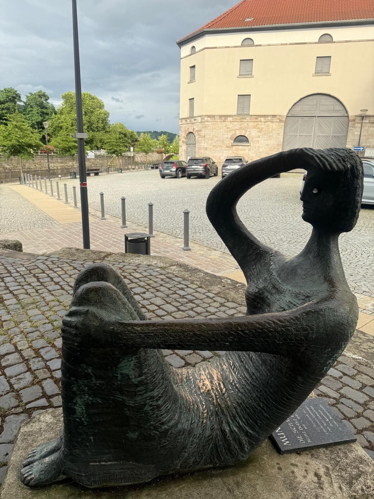 Une sculpture en bronze représentant une figure allongée sur un chemin pavé, avec un bâtiment en arrière-plan.