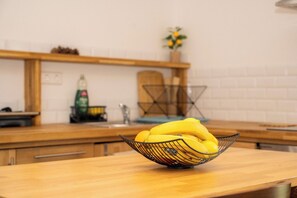 A kitchen with a wooden countertop, a metal fruit basket containing bananas and an orange, a sink, and a plant in the background.