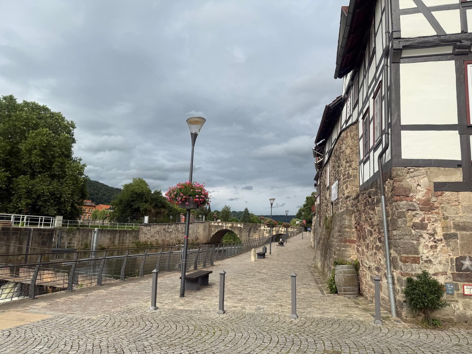 Un sentiero in pietra ciottolata conduce a un ponte di pietra su un fiume, con un edificio storico su un lato e alberi sullo sfondo.