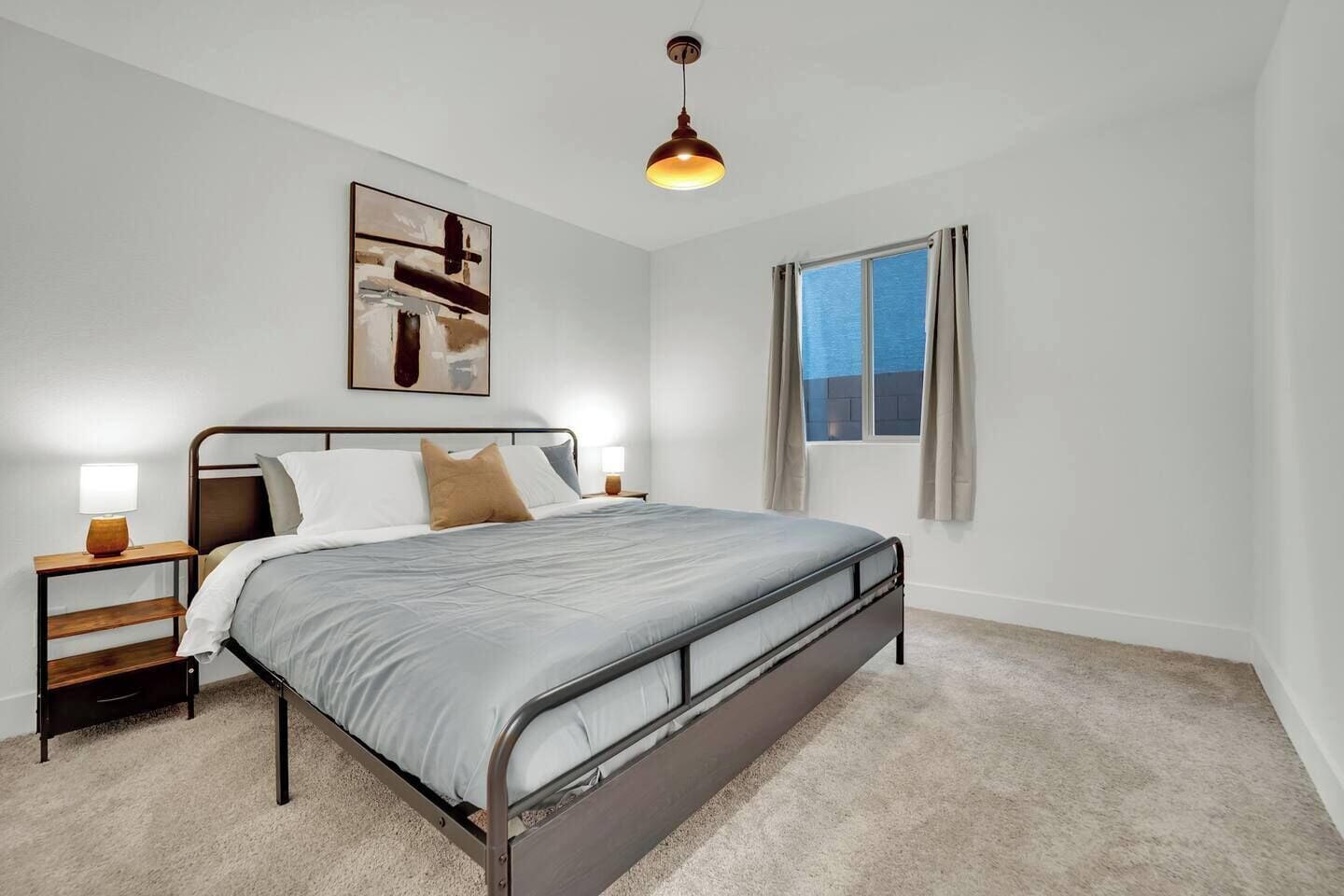 Quarto com estrutura de cama em metal, mesa de cabeceira com abajur, obra de arte na parede e janela com cortinas.