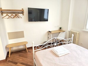 A bedroom with a bed, a TV mounted on the wall, a wooden bench, a small table with a plant, and a radiator. - DOUBLE ROOM WITH PRIVATE BATHROOM (Milano)