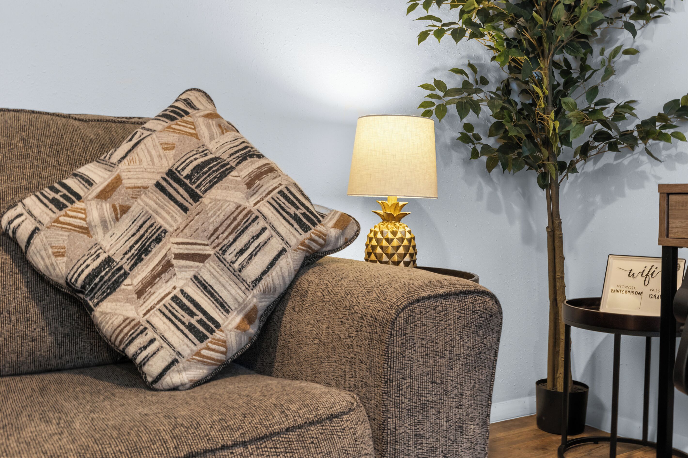 Ein Sofa mit einem gemusterten Zierkissen, eine Lampe mit geometrischem Fuß und eine Topfpflanze an einer Wand.