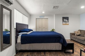 Een slaapkamer met een bed, een spiegel, een deur, een raam met jaloezieën, een wanddecoratie en een bank.
