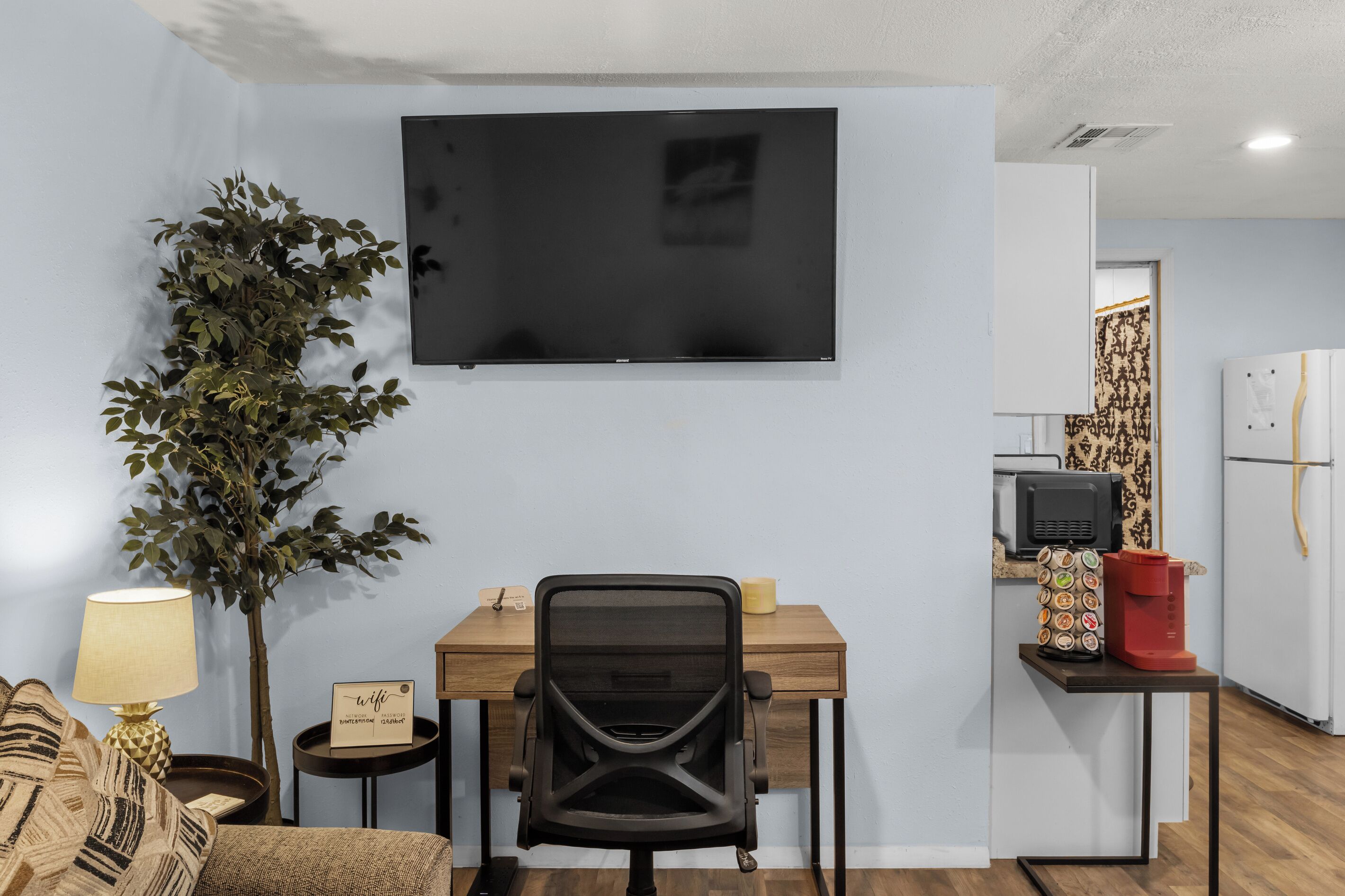 A room with a TV mounted on the wall, a desk with a chair, a small table with a lamp, a potted plant, and a kitchen area with a refrigerator and a coffee machine.