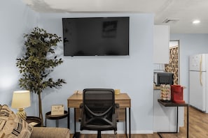 A room with a TV mounted on the wall, a desk with a chair, a small table with a lamp, a potted plant, and a kitchen area with a refrigerator and a coffee machine.