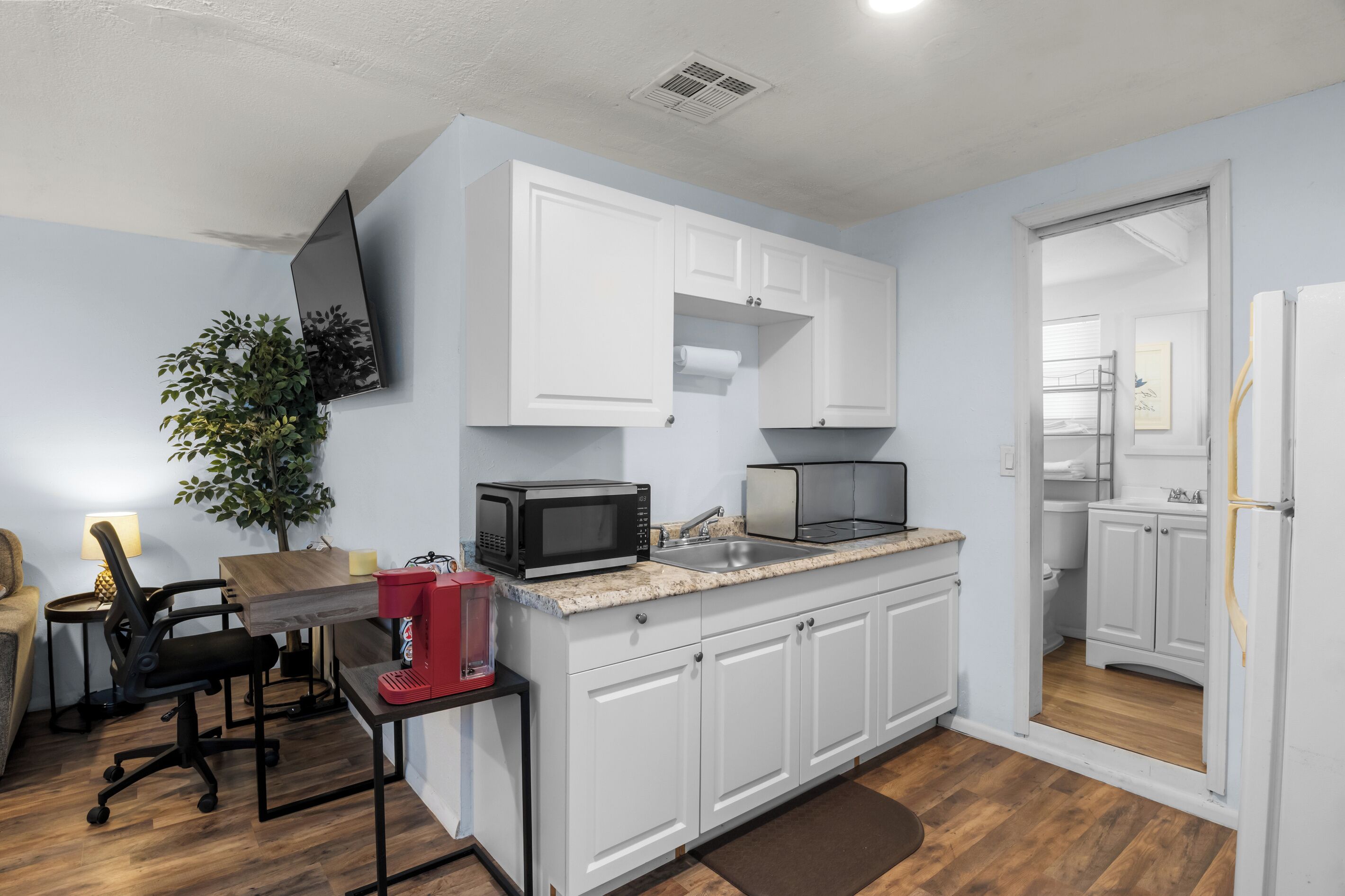 A compact kitchen with white cabinets, a microwave, a sink, and a small dining area with a chair and table.