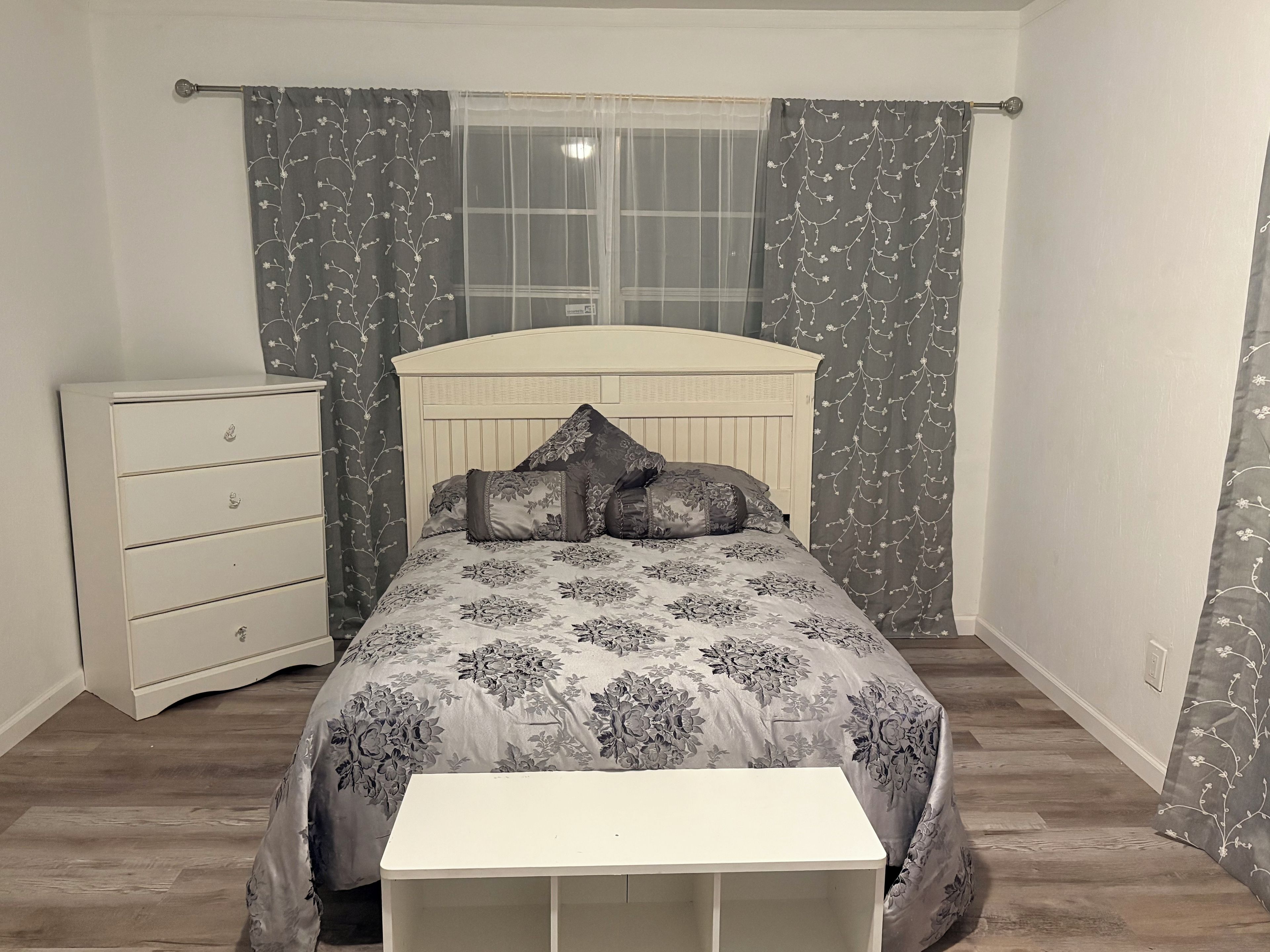 A bedroom with a bed, a bedside table, a dresser, and a window with curtains.