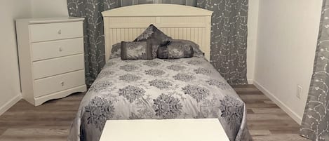 Une chambre à coucher avec un lit, une table de chevet, une commode et une fenêtre avec des rideaux.