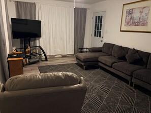 A living room with a sectional sofa, a leather armchair, a flat-screen TV, a small side table, and a patterned rug. - Charming & Spacious In-Town Oasis – Mins to Springs, Dining & Beaches! (Crystal River)