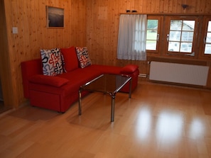 Ein Zimmer mit einem roten Sofa, einem Glastisch, Holzwänden, einem Fenster mit Vorhängen und einem Heizkörper.