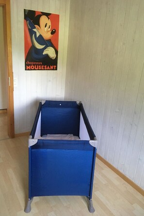 Ein Zimmer mit einem blauen Reisebett, einer holzverkleideten Wand, einem Poster mit einer Zeichentrickfigur und einer Holztür.