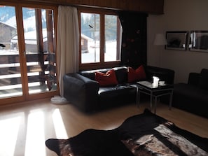 Uma sala de estar com um sofá, uma mesa de centro e um tapete. Há uma porta de vidro deslizante que dá para uma varanda.