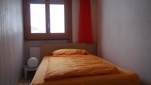Uma cama de solteiro com um edredom laranja num quarto com paredes de madeira e uma janela com cortina vermelha.