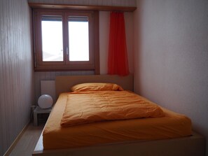 Ein Einzelbett mit orangefarbener Bettdecke in einem Zimmer mit Holzwänden und einem Fenster mit rotem Vorhang.