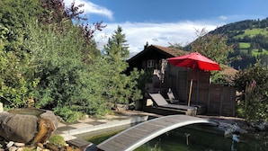 Une terrasse en bois mène à un étang surmonté d’un petit pont, le tout étant entouré de verdure et ayant une montagne en toile de fond.