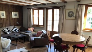 Un salon chaleureux avec un canapé, une table basse et une table à manger avec des chaises. De grandes fenêtres sont ornées de rideaux, le plafond est en bois et un tableau encadré est accroché au mur.