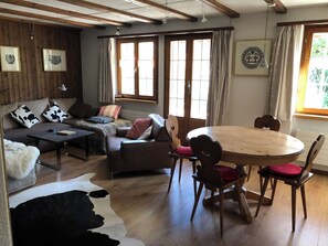 A cozy living room with a sofa, a coffee table, and a dining table with chairs. There are large windows with curtains, a wooden ceiling, and a framed picture on the wall. - Strubeli Appartement/Fewo, Dusche oder Bad, WC, 2 Schlafräume 4-Bett-Wohnung by Interhome (St Stephan)