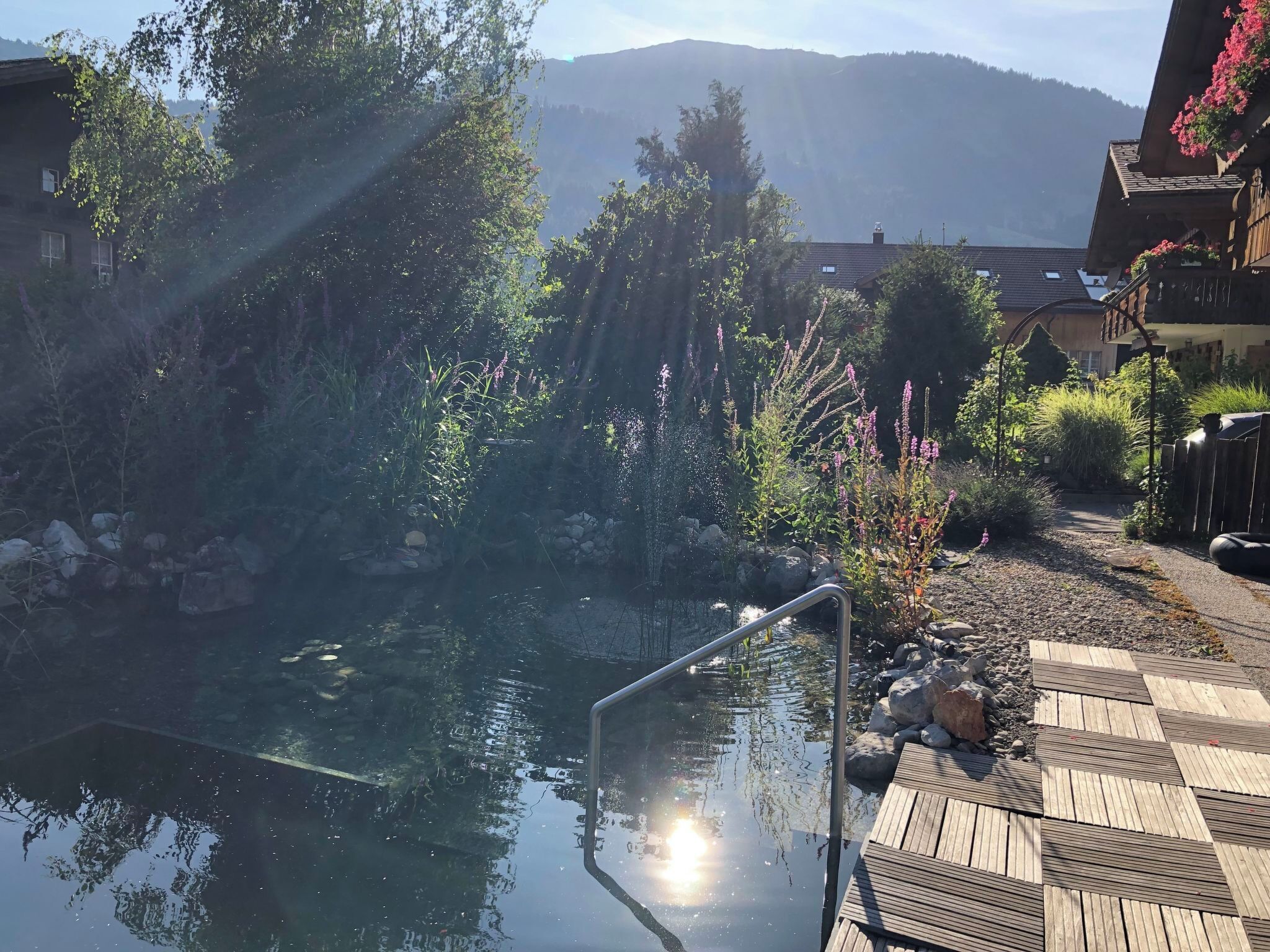 Una piscina esterna trasparente con un solarium in legno, immersa nel verde con montagne sullo sfondo.