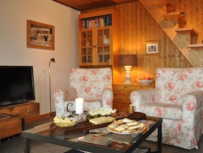 Ein gemütliches Wohnzimmer mit stanzmuster-gepolsterten Sesseln, einem Couchtisch mit Snacks, einem Fernseher und einem Bücherregal.