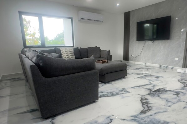 A modern living room with a grey sofa set, a flat-screen TV mounted on the wall, and a large window providing natural light.