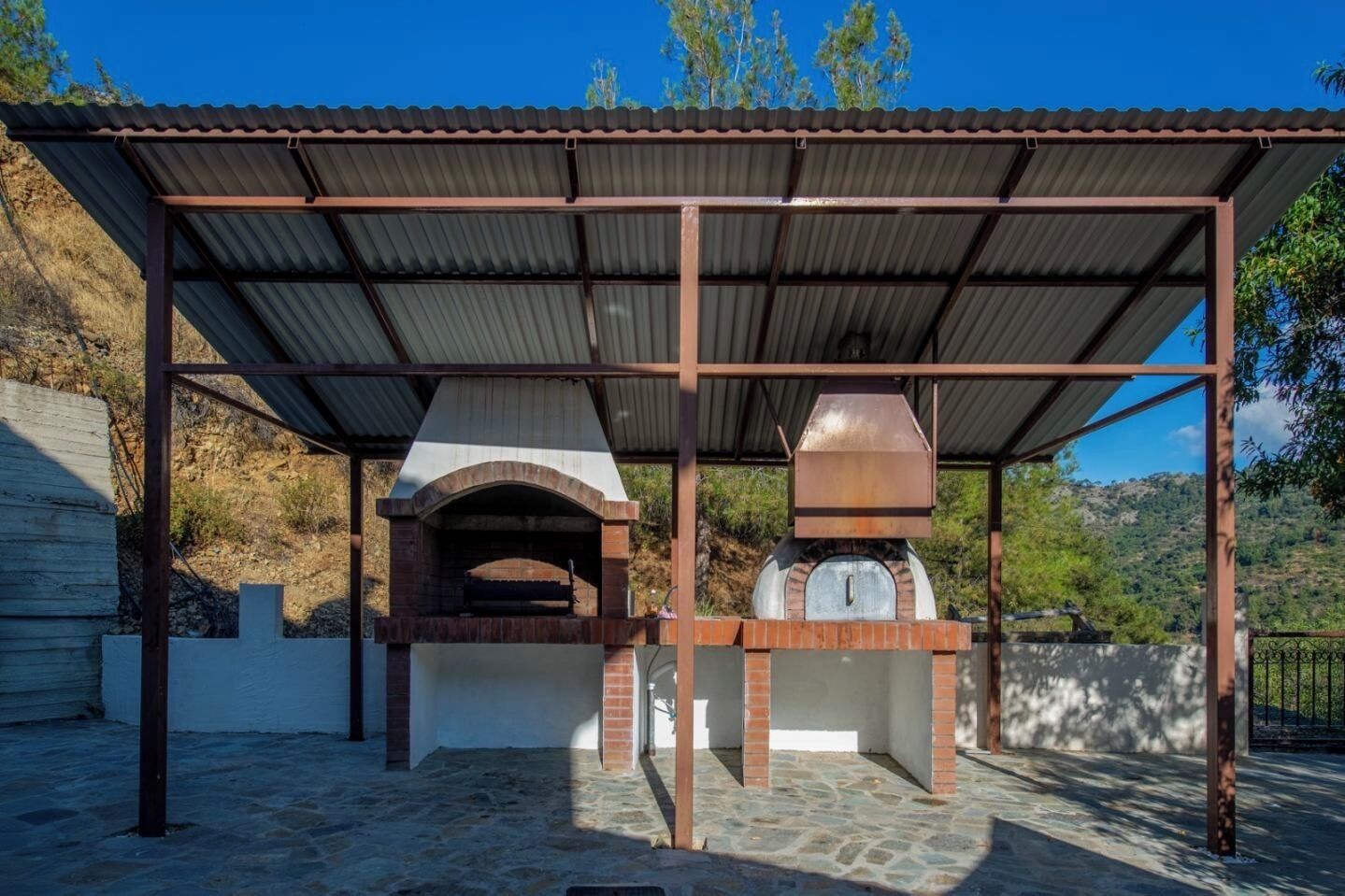 An outdoor brick oven structure with a metal roof, surrounded by a stone-paved area.