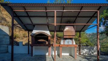 An outdoor brick oven structure with a metal roof, surrounded by a stone-paved area.
