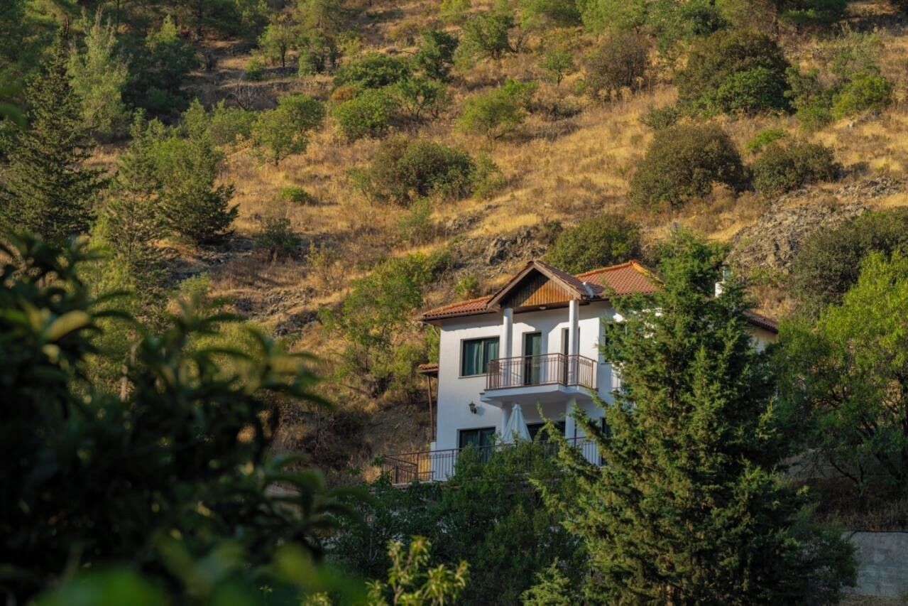 Une maison à deux étages avec un balcon, entourée d’arbres et de collines.
