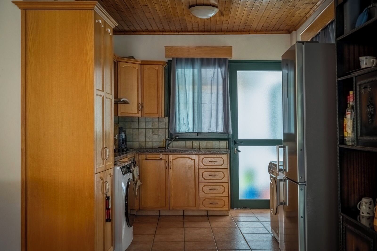Une cuisine compacte avec des meubles en bois, un lave-linge encastré et une grande fenêtre avec des rideaux.