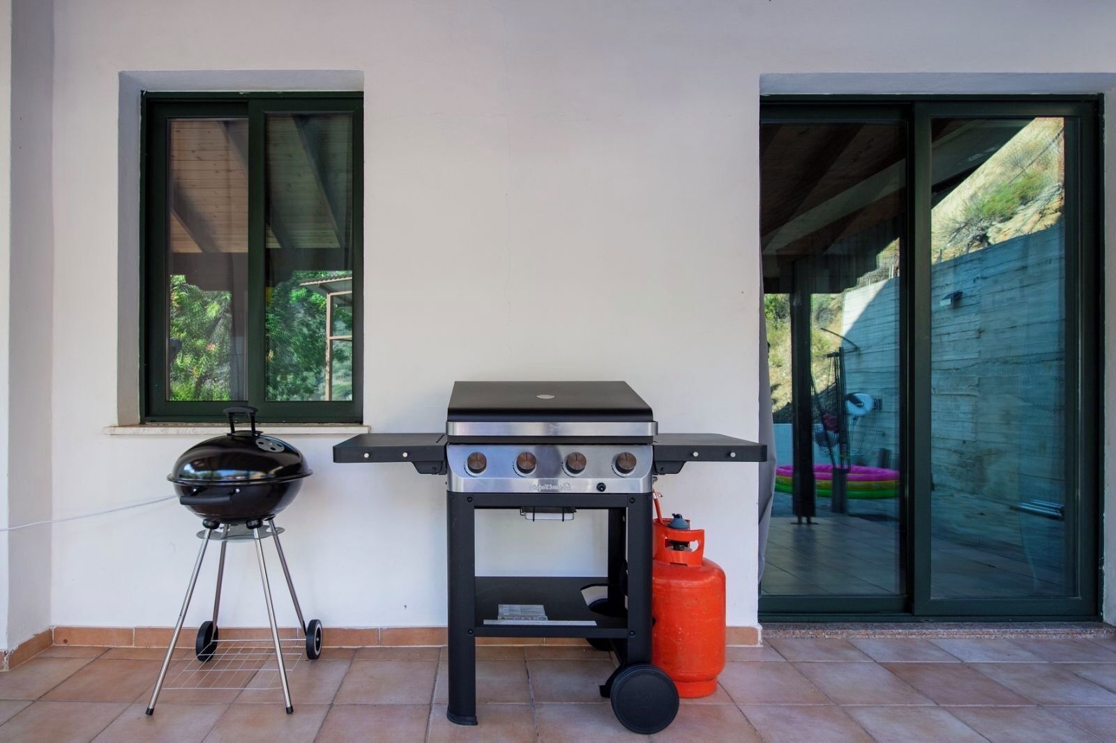 Une terrasse extérieure équipée d’un barbecue, d’une bouteille de gaz et d’une fenêtre donnant sur un paysage verdoyant.