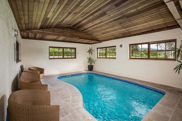 An indoor swimming pool with a wooden ceiling, clear blue water, and wicker chairs. - Wahroonga Luxury Estate in Kangaroo Valley (Kangaroo Valley)