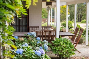 An outdoor patio with a wooden table and chairs, surrounded by lush greenery and hydrangea flowers. - Wahroonga Luxury Estate in Kangaroo Valley (Kangaroo Valley)