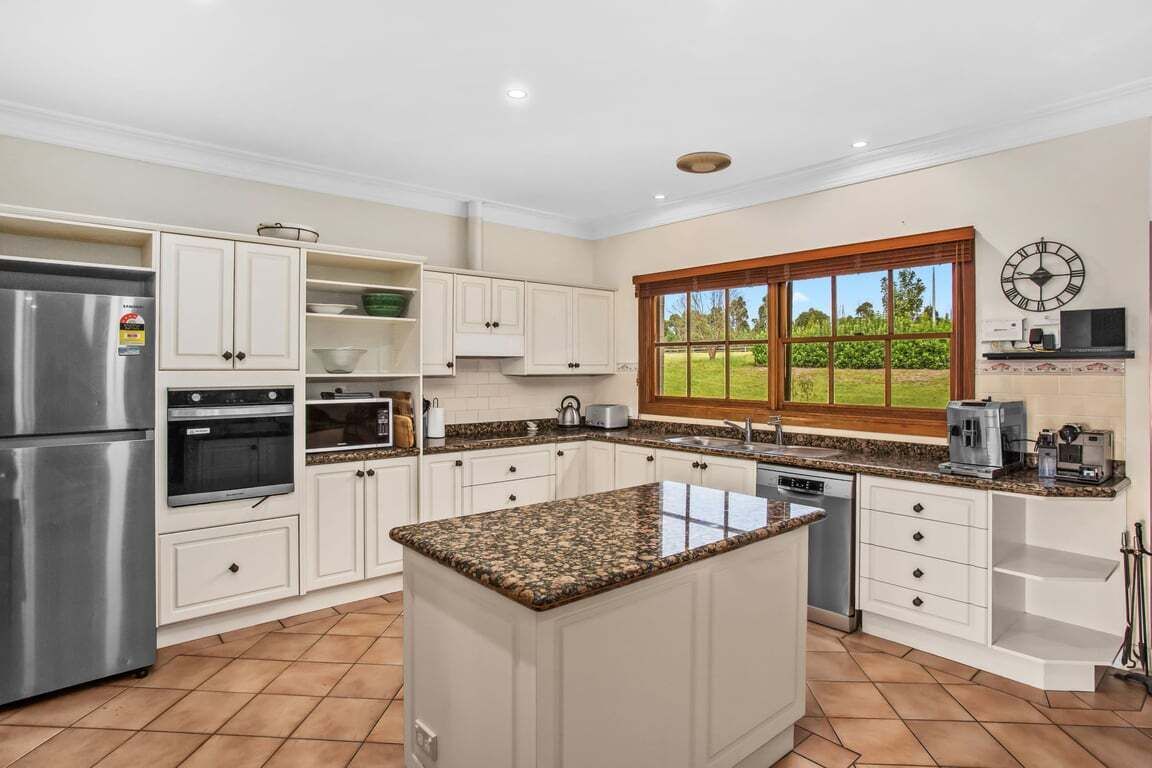 A kitchen with white cabinetry, a granite countertop island, a stainless steel refrigerator, and a window with a view of greenery.