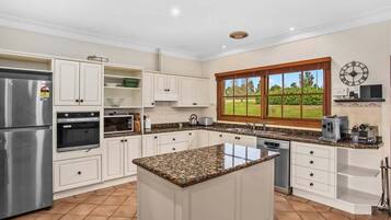 A kitchen with white cabinetry, a granite countertop island, a stainless steel refrigerator, and a window with a view of greenery.