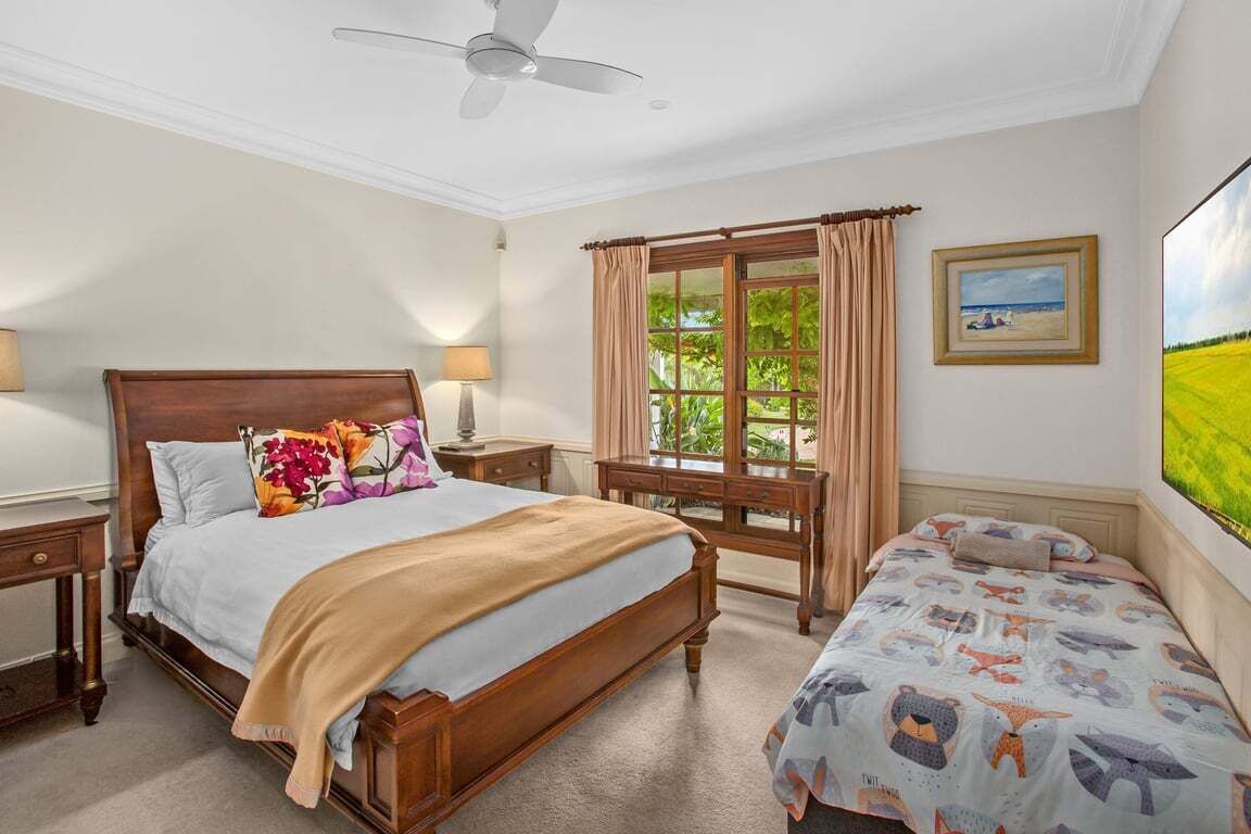A bedroom with a large bed, a smaller bed, a desk, a chair, a ceiling fan, and a window with curtains.