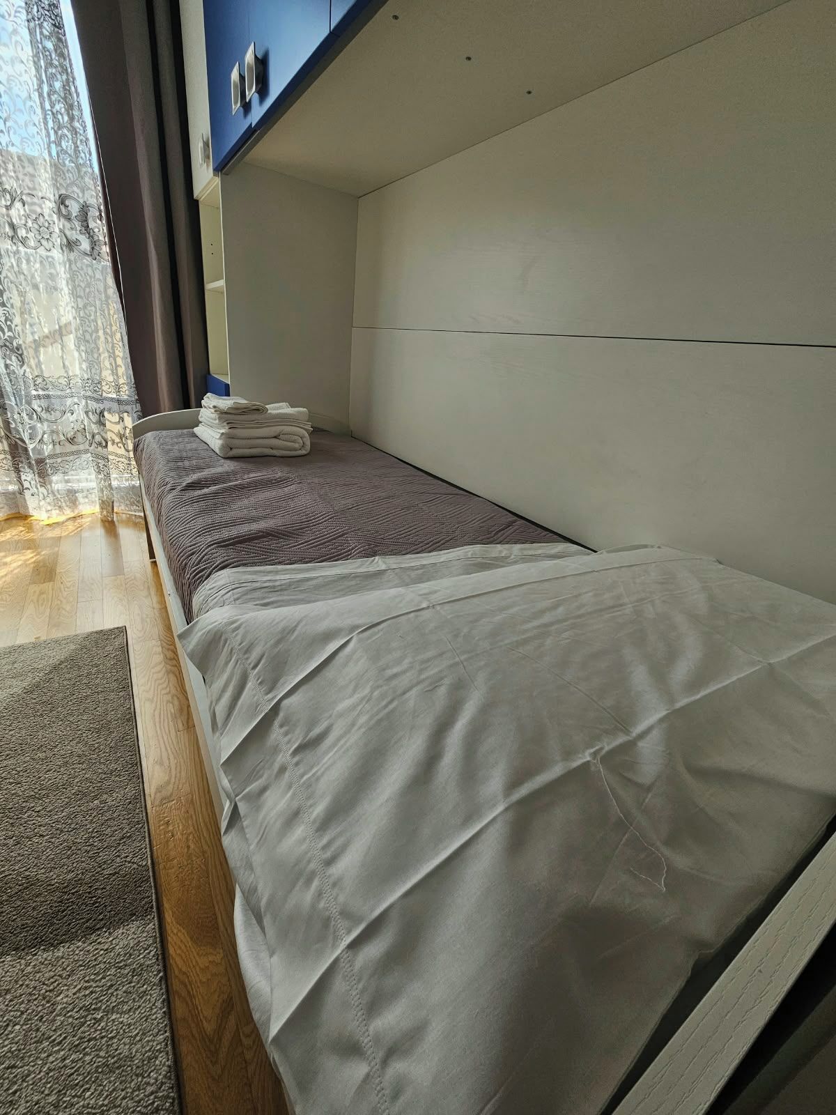 A hotel room with a bed, a window with curtains, a wall-mounted control panel, and a small stack of folded towels.