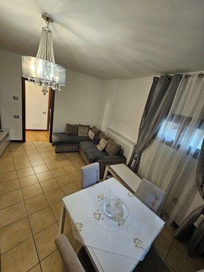 Un salon avec un canapé, une table à manger entourée de chaises, un lustre et une vue à travers les rideaux.